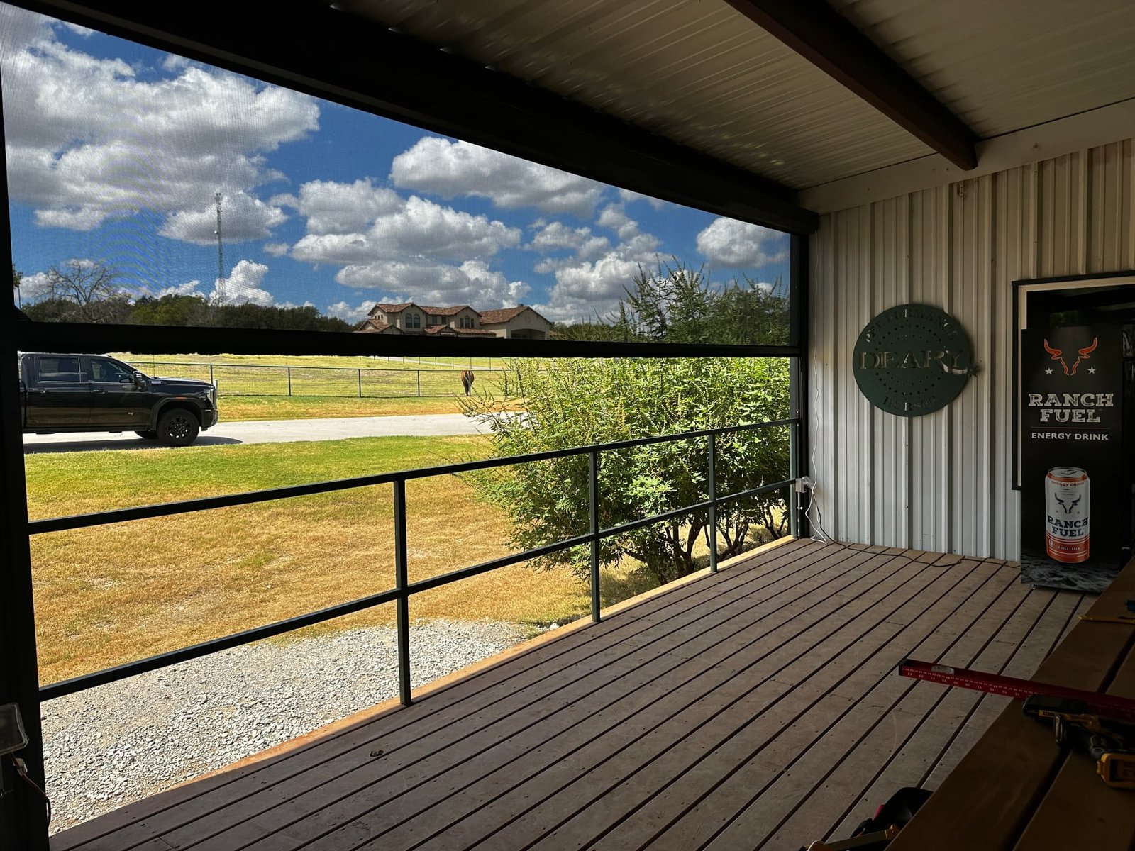 A covered ranch deck with railing overlooking pasture, truck, horse, and distant home under a wide Texas sky.